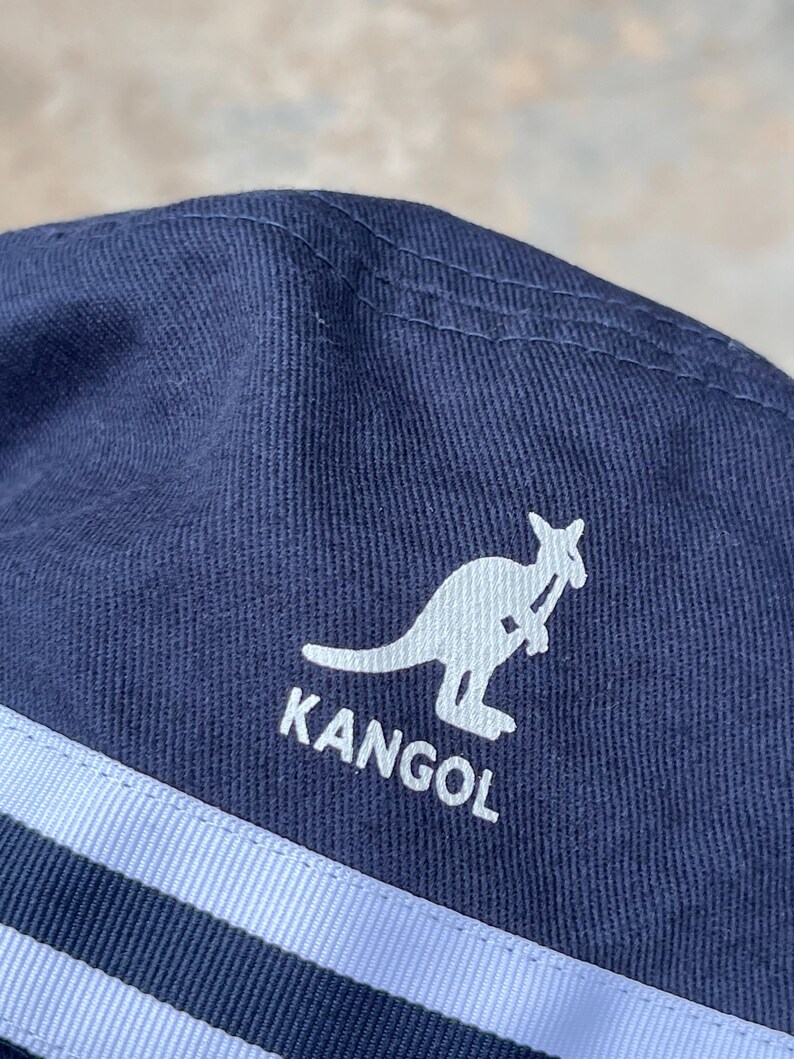 May include: Navy blue Kangol hat with a white kangaroo logo and the brand name. The hat features two light blue stripes and is made of a textured fabric. The hat is a classic style.