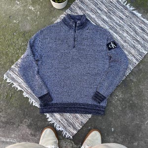 May include: A navy blue and white speckled knit quarter-zip jumper featuring a high collar and a black patch on the left sleeve with the white text CK. The jumper has ribbed cuffs and a ribbed hem in a darker solid navy blue tone.