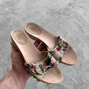 May include: A pair of Gucci clogs with a floral print and gold-tone hardware. The wooden soles are light brown, and the straps feature a colorful floral pattern with the Gucci logo.