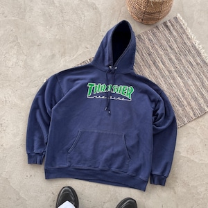 May include: A navy blue hoodie with a front pocket and a drawstring hood. The front displays the text "THRASHER MAGAZINE" in green and white. The hoodie is made of a soft material.