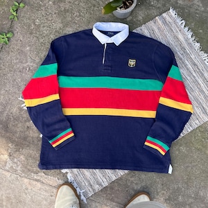 May include: A navy blue long-sleeve rugby shirt featuring a white collar and a button placket. The chest and sleeves are decorated with horizontal stripes in green, red, and yellow. A small embroidered crest logo is positioned on the left chest.