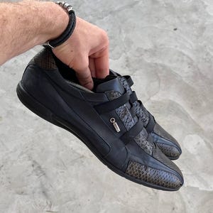 Może przedstawiać: Czarne skórzane sneakersy z akcentami wężowej skóry. Buty mają niski profil, pasek na górze i mały metalowy detal. Osoba trzyma czarną skórzaną bransoletkę.