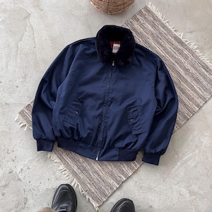 May include: A navy blue bomber jacket with a black faux fur collar. The jacket has a zipper closure, two front pockets, and ribbed cuffs and hem. The jacket is lying on a woven rug.