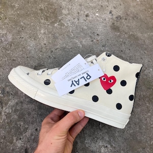 May include: A high-top sneaker in cream canvas with black polka dots, featuring a red heart logo with eyes on the side. A white tag attached to the laces displays the text PLAY COMME des GARÇONS in black ink. The shoe has a white rubber sole.