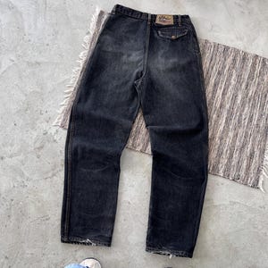 May include: Dark wash denim jeans with a faded appearance. The jeans have a classic five-pocket design, a button and zipper closure, and a leather patch on the back. The jeans are laid flat on a textured surface.