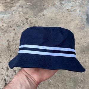 May include: Navy blue bucket hat with two white stripes. The hat has a classic design with a slightly downward-sloping brim. The hat is made of a sturdy fabric, suitable for sun protection and casual wear.
