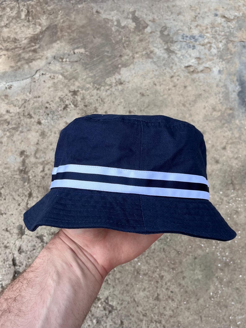 May include: Navy blue bucket hat with two white stripes. The hat has a classic design with a slightly downward-sloping brim. The hat is made of a sturdy fabric, suitable for sun protection and casual wear.