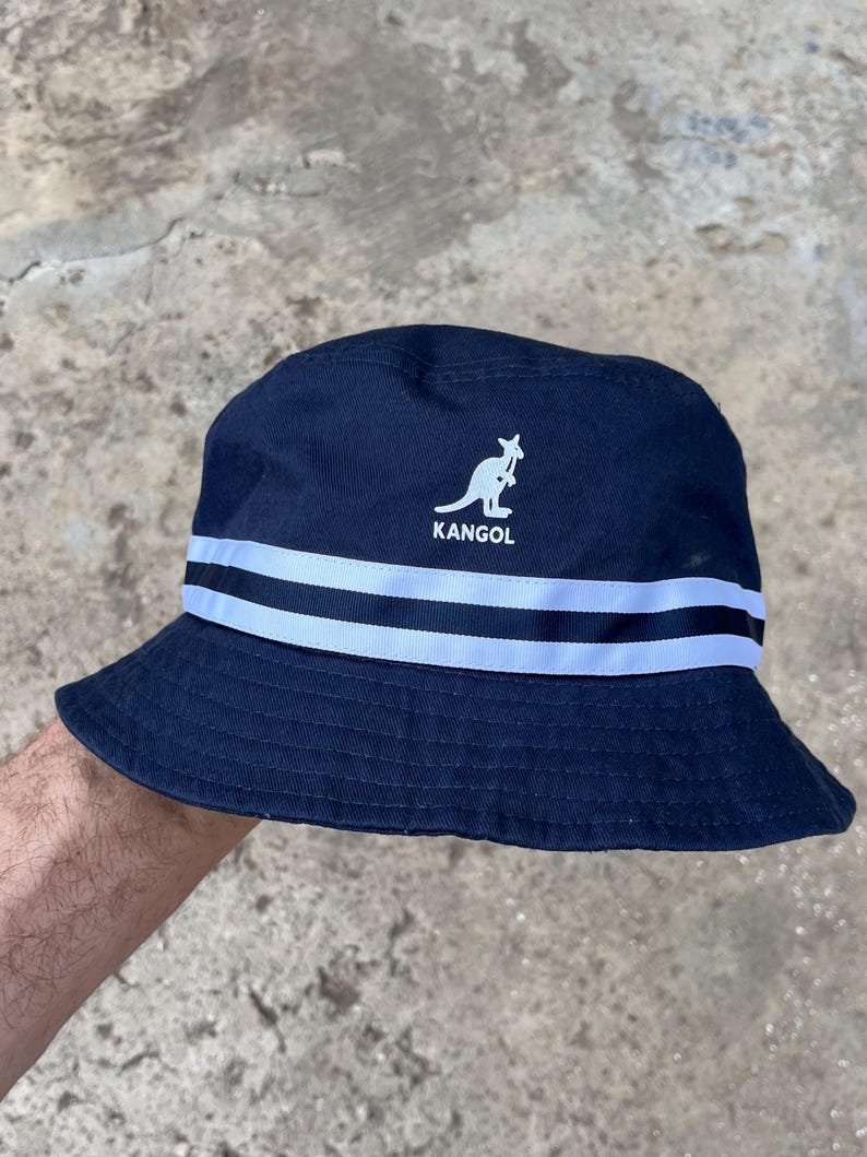 May include: Navy blue bucket hat with white stripes and the word "KANGOL" below a kangaroo logo. The hat is made of a soft material and is suitable for casual wear. The hat is a classic style.