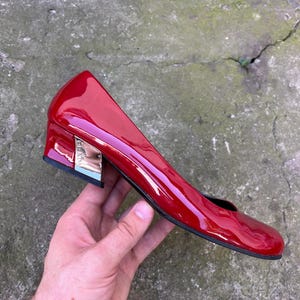 May include: A glossy red patent leather pump with a rounded toe and a low, block-shaped heel. The side of the metallic silver heel features the Prada logo embossed in a vertical orientation. The shoe has a black sole and a smooth, reflective finish.