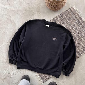 May include: Black crew neck sweatshirt featuring the Nike logo in silver embroidery. The long-sleeved top is made of a soft fabric. The image shows the sweatshirt on a neutral background.