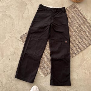 May include: Black work pants with reinforced knee panels. The pants have a straight leg design and a small logo patch on one knee. The pants are laid flat on a patterned rug, with a woven basket and a pair of white sneakers in the background.