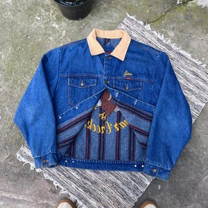 May include: A blue denim jacket with a tan corduroy collar and long sleeves. The interior features a dark blue, red, and grey striped lining with a large embroidered graphic of a brown eagle and the yellow text "Joe's Creek" on the back panel.