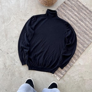 May include: A black turtleneck jumper with long sleeves. The jumper is made of a soft material and has a ribbed collar and cuffs. The jumper is lying on a grey surface next to a woven rug and a basket with greenery.