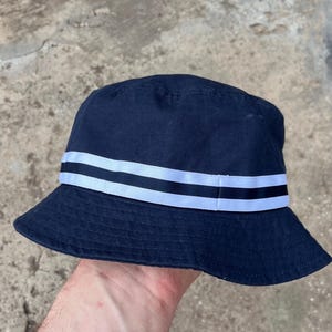 May include: Navy blue bucket hat with a white and black striped band around the crown. The hat is made of a soft fabric and has a classic, casual style. The hat is held by a person's hand.