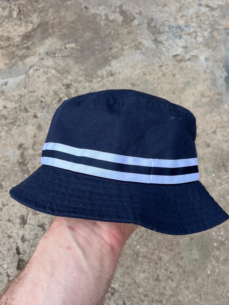 May include: Navy blue bucket hat with a white and black striped band around the crown. The hat is made of a soft fabric and has a classic, casual style. The hat is held by a person's hand.