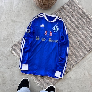 以下が含まれることがあります： 白のストライプとアクセントが付いた、ロイヤルブルーの長袖サッカーユニフォーム。ユニフォームにはアディダスのロゴ、チームエンブレム、キリル文字が描かれています。ストライプのラグの上にユニフォームが置かれています。