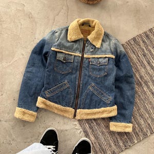 May include: A blue denim jacket with a tan faux fur collar and trim. The jacket has two chest pockets and two side pockets. It features a zipper closure and a red tag on the chest pocket. The jacket is a vintage style.