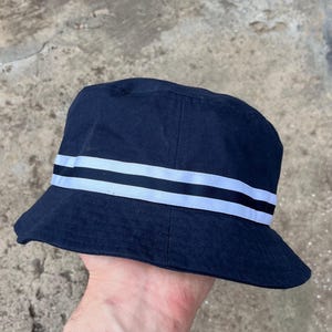 May include: Navy blue bucket hat with two white stripes and a black stripe. This casual hat is made of a durable fabric and is suitable for various outdoor activities. The hat's brim provides sun protection.
