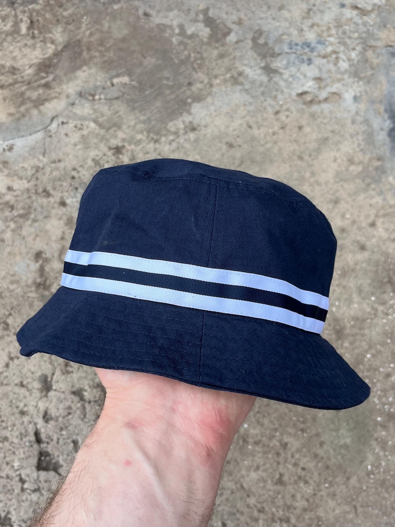 May include: Navy blue bucket hat with two white stripes and a black stripe. This casual hat is made of a durable fabric and is suitable for various outdoor activities. The hat's brim provides sun protection.