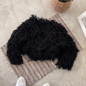 Puede incluir: Una chaqueta negra de punto peludo con una silueta recortada. La chaqueta está hecha de hilo grueso y texturizado, creando una apariencia densa y con flecos. Se exhibe sobre una alfombra a rayas, con una cesta tejida de vegetación en el fondo.
