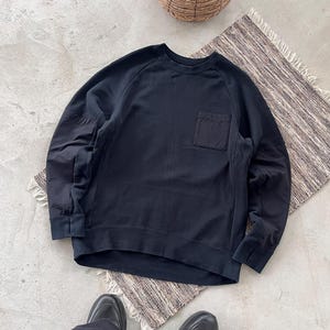 May include: A black crew neck sweatshirt with a single pocket on the chest. The long-sleeved top is made of a soft material and is shown on a neutral-coloured rug.
