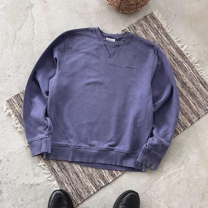 May include: A periwinkle blue sweatshirt with the brand name "Carhartt" embroidered on the chest. The crew neck sweatshirt is on a patterned rug with a woven basket and plant in the background. Black shoes and camouflage pants are visible.