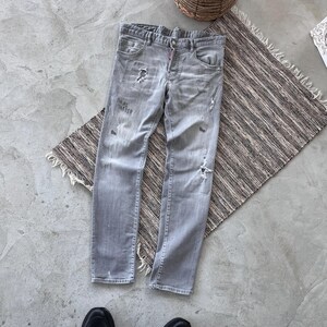 Puede incluir: Vaqueros de mezclilla gris claro con detalles desgastados, incluyendo rasgaduras y parches descoloridos. Los vaqueros tienen un diseño clásico de cinco bolsillos y bragueta con botones. El texto "TAKE MATTER" es visible en la parte delantera. Los vaqueros están sobre una alfombra a rayas.