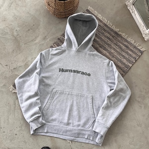 May include: A light grey hoodie featuring the word "Humanrace" printed on the front. The hoodie has a kangaroo pocket and a hood. The image is taken from above, with a plant in a woven basket and a mirror in the background.