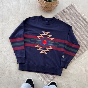 Puede incluir: Sudadera azul marino con un diseño geométrico en rojo, verde y beige. La sudadera tiene puños acanalados y cuello redondo. El logotipo de la marca es visible en la manga.