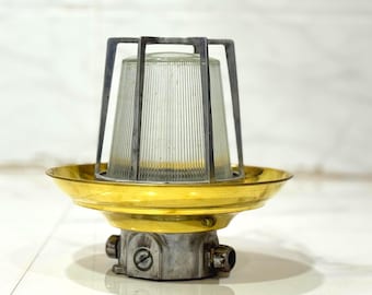 Peut inclure: Un luminaire de style industriel avec un abat-jour en verre transparent et nervuré, protégé par une cage en métal sombre. Il possède une base de couleur laiton et un boîtier en métal gris avec des composants de câblage visibles. Design vintage ou nautique.
