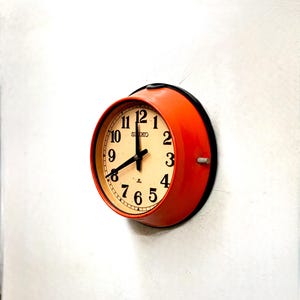 Puede incluir: Un reloj de pared Seiko redondo, naranja, con una esfera color crema y números negros. El reloj está montado en una pared blanca. Las manecillas del reloj señalan aproximadamente las 8:00.