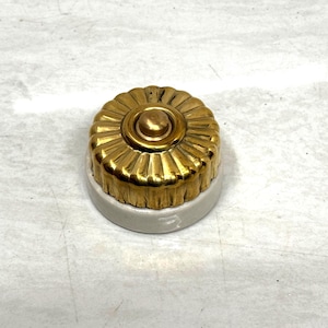 May include: A vintage doorbell button with a gold-coloured, fluted face and a small, circular button in the centre. The base is a light grey colour. The doorbell button is likely made of metal and ceramic.