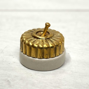 May include: A vintage-style light switch with a gold-colored, fluted cover and a small, matching toggle. The switch sits on a light gray, circular base. The design is reminiscent of antique electrical fixtures.