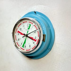 Könnte beinhalten: Eine Vintage Citizen Wanduhr mit hellblauem Metallgehäuse. Das Zifferblatt ist weiß mit schwarzen Ziffern, roten und grünen Akzenten und schwarzen Zeigern. Die Uhr ist an einer weißen Wand befestigt.