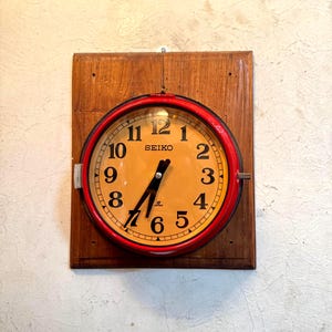 May include: A vintage Seiko wall clock with a red bezel and a cream-colored face. The clock is mounted on a wooden backing. The clock hands are black, and the numbers are black. The clock is hanging on a textured, off-white wall.
