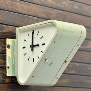 May include: A double-sided wall-mounted Seiko clock with a pale green, trapezoidal metal housing. The clock face is white with black hour markers and black hands. The mounting bracket shows signs of chipped paint, and the casing has surface scratches.