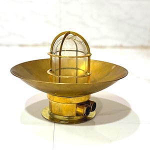 Puede incluir: Una lámpara náutica vintage de latón. Presenta un globo de cristal transparente encerrado en una jaula protectora, montado sobre una base circular con un plato acampanado. El latón tiene una pátina cálida y envejecida.
