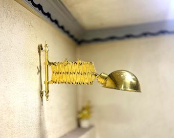 May include: A brass-colored, articulated wall-mounted lamp. The lamp features a concertina-style arm and a rounded shade. It is mounted on a textured, off-white wall. The lamp is designed to be adjustable and directional.
