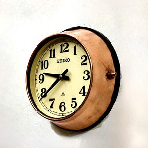 Könnte beinhalten: Eine Vintage Seiko Wanduhr mit kupferfarbenem Rahmen und cremefarbenem Zifferblatt. Die Uhr hat schwarze Zeiger und Ziffern und ist an einer weißen Wand befestigt. Das Zifferblatt zeigt "SEIKO".