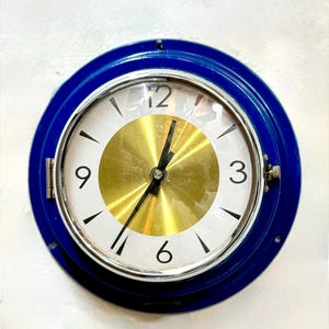 May include: A round wall clock with a blue metal frame. The clock face is white with black numerals and hands, and a gold-colored center. The clock is mounted on a white wall.
