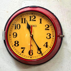 May include: A round wall clock with a red frame and a yellow face. The clock features black numerals and hands, with the brand name "SEIKO" visible. The clock is mounted on a textured white wall.
