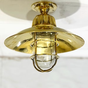 Może przedstawiać: Mosiężna lampa sufitowa z zaokrąglonym kloszem i przezroczystą szklaną obudową. Światło ma polerowane wykończenie i konstrukcję przypominającą klatkę wokół szkła. Oprawa jest zamontowana na białym suficie.