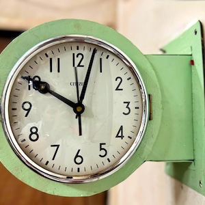 Op de afbeelding: Een ronde, lichtgroene Citizen-wandklok met een witte wijzerplaat, zwarte cijfers en zwarte wijzers. De klok is voorzien van een metalen zilverkleurige rand en een bijpassende lichtgroene montagebeugel die aan de zijkant is bevestigd.