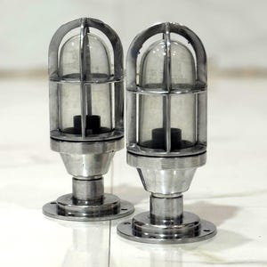May include: Two industrial-style, silver-toned metal lamps. Each lamp features a clear, rounded glass enclosure protected by a metal cage. The lamps are mounted on a circular base with a central post.