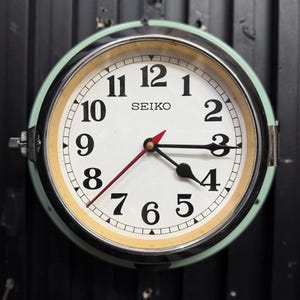 Puede incluir: Un reloj de pared redondo Seiko con esfera blanca, números arábigos negros y una carcasa exterior de color verde claro. El reloj cuenta con manecillas negras para horas y minutos, un segundero rojo fino y un bisel de metal cromado.