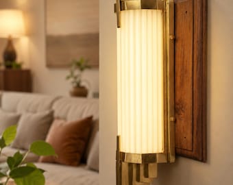 Peut inclure: Applique murale de style Art déco avec une base en bois rectangulaire. L'applique présente un luminaire en verre cannelé vertical, encadré dans un cadre en laiton. Le cadre en laiton présente des détails géométriques étagés. La lumière est allumée.