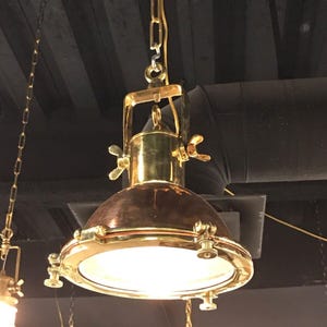 May include: A vintage-style brass and copper pendant light fixture. The light has a rounded, copper-colored shade with a brass rim and accents. It is suspended from a gold chain and features wingnut details. The light is turned on, illuminating the room.