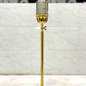 Puede incluir: Una lámpara alta de color dorado con una pantalla en forma de jaula. La lámpara tiene una carcasa de vidrio cilíndrica y una base decorativa. La lámpara está hecha de latón y tiene un diseño vintage e industrial. La lámpara está sobre un suelo de mármol.