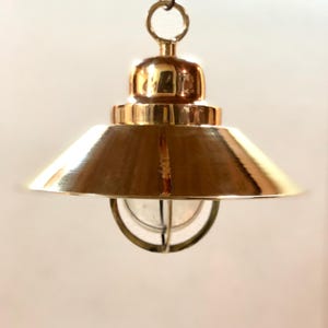 Puede incluir: Una lámpara colgante de color latón con una pantalla cónica y una jaula protectora de alambre alrededor de la bombilla de vidrio. La lámpara se suspende de un pequeño anillo.