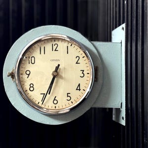 May include: A vintage Citizen clock with a light blue frame and a silver-colored bezel. The clock face is cream-colored with black numerals and hands. The clock is mounted on a dark surface.
